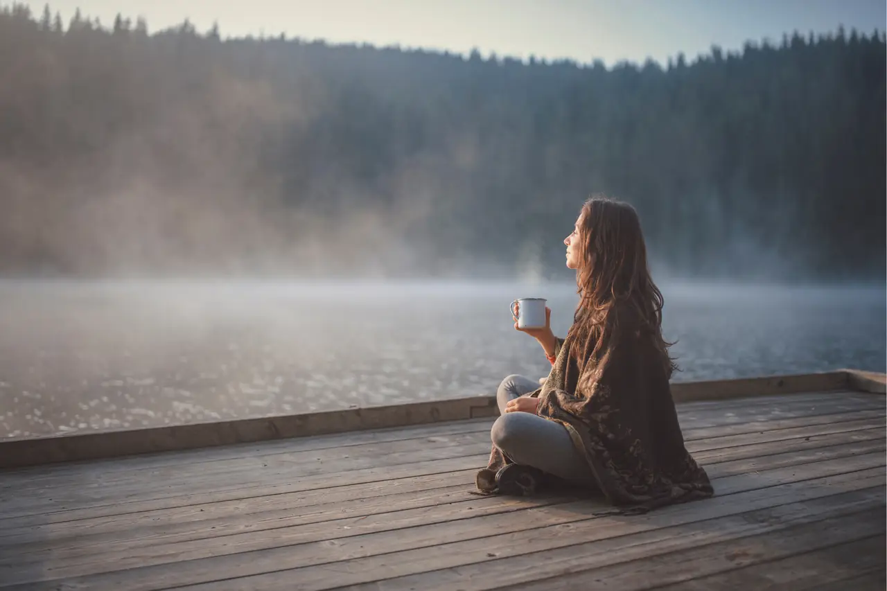 Eine Frau sitzt mit einer Tasse in der Hand auf einem Steg an einem ruhigen See
