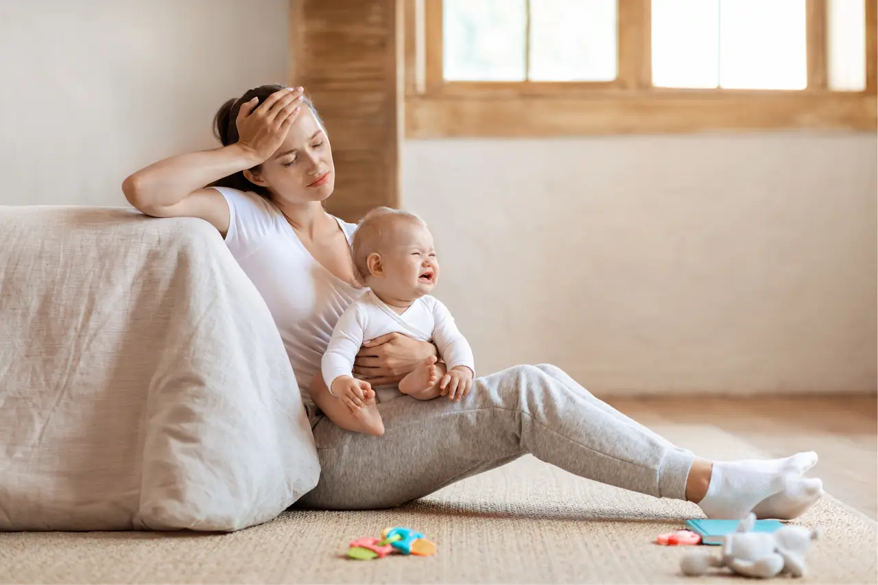 Eine erschöpfte Mutter sitzt mit ihrem Baby auf dem Boden, die Hand an der Stirn